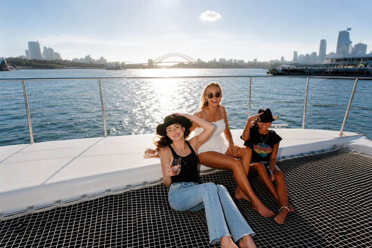 Three friends relaxing on the bow net of a charter yacht with the Sydney Harbour Bridge behind
