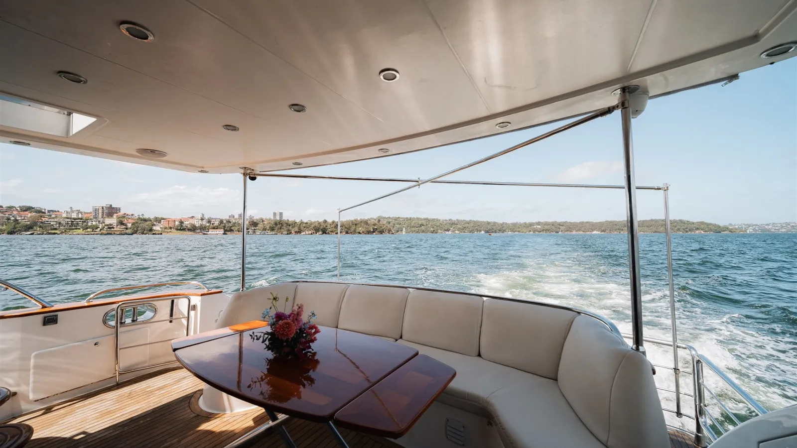 Enigma's covered aft deck with lounge seating and a timber table, cruising on the harbour